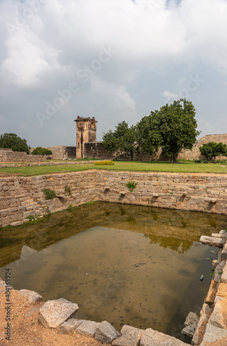 Hampi, Karnataka, India - November 5, 2013: Zanana Enclosure. Closeup of tank or pool up front and brown stone watchtower and wall in back under blue cloudscape. Green foliage and grass. photo