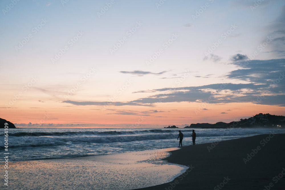 Fototapeta premium Atardecer en playa de Ixtapa Zihuatanejo, Mexico, atardecer en tono naranja y azul con un par de siluetas sobre la arena y cerca del mar. 