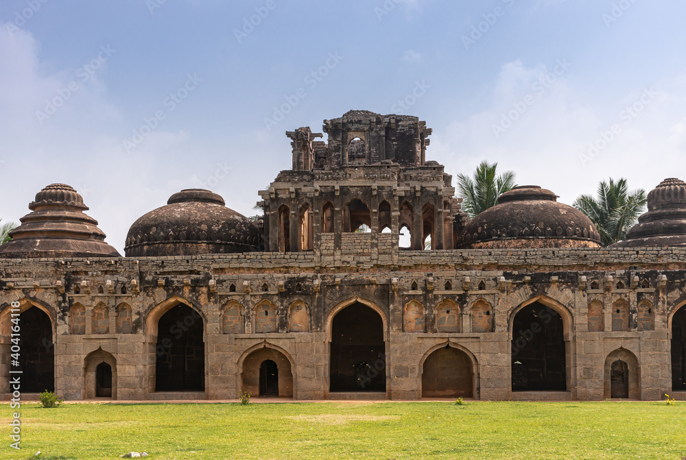 Hampi, Karnataka, India - November 5, 2013: Zanana Enclosure. Central part of Brown stone ruinous Elephant stables with multiple domes seen over green lawn under blue cloudscape. 