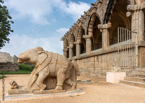 Hampi, Karnataka, India - November 5, 2013: Zanana Enclosure. Brown stone elephant statue in front of archeological items exhibition hall under blue cloudscape with green foliage. photo