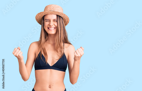 Young beautiful girl wearing bikini and hat very happy and excited doing winner gesture with arms raised, smiling and screaming for success. celebration concept.