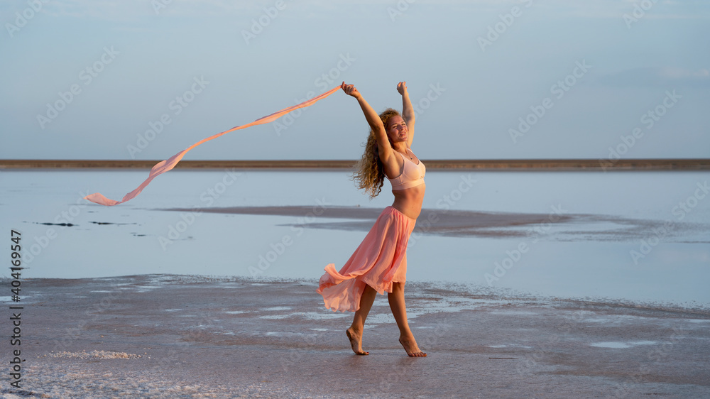 beautiful graceful girl in the rays of pink sunset light on the seashore keeps a pareo developing in the wind. lightness, freedom