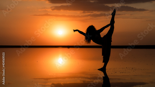silhouette of a flexible girl performing split on the background of a golden sunset mirrored reflection in the water
