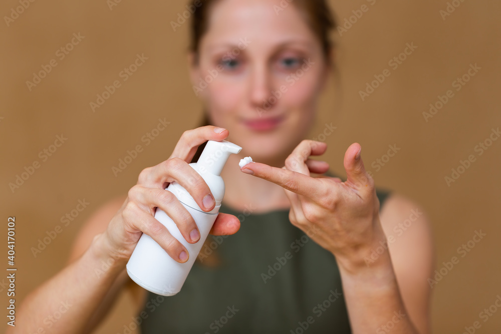 Happy young woman pushing white pump and applying white cream on her fingers. Female using moisturizer on her skin. Beauty cosmetic product in use with blurred face in background.