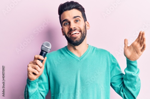 Young handsome man with beard singing song using microphone celebrating achievement with happy smile and winner expression with raised hand