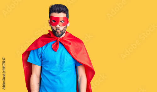 Foto Young handsome man with beard wearing super hero costume making fish face with lips, crazy and comical gesture