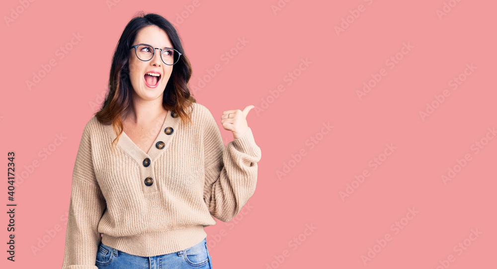 Beautiful young brunette woman wearing casual clothes and glasses smiling with happy face looking and pointing to the side with thumb up. Beautiful young brunette woman wearing casual clothes and glasses smiling with happy face looking and pointing to the side with thumb up.
