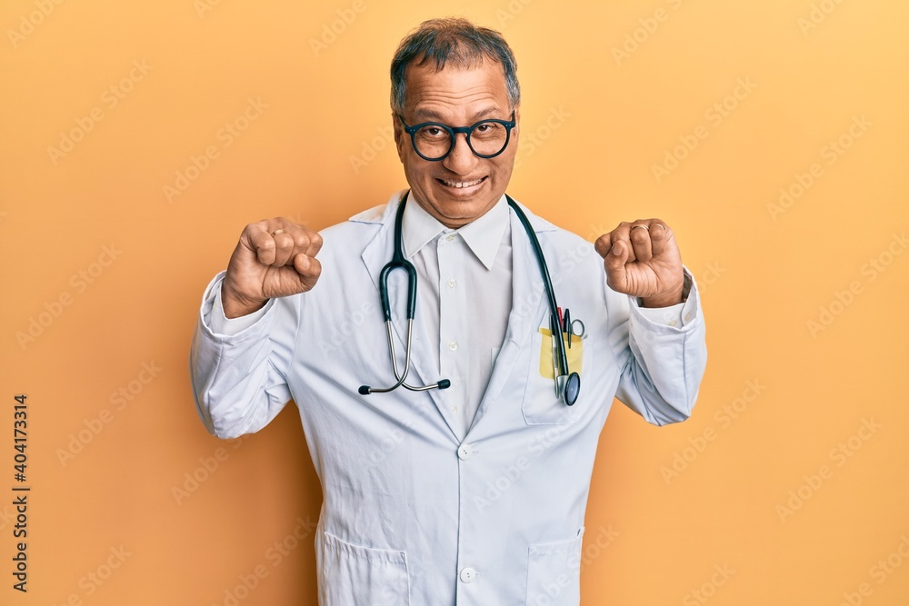 Middle age indian man wearing doctor coat and stethoscope celebrating surprised and amazed for success with arms raised and open eyes. winner concept. Middle age indian man wearing doctor coat and stethoscope celebrating surprised and amazed for success with arms raised and open eyes. winner concept.
