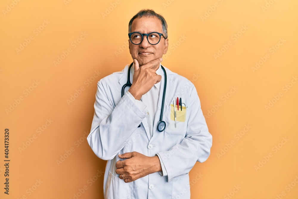 Middle age indian man wearing doctor coat and stethoscope with hand on chin thinking about question, pensive expression. smiling with thoughtful face. doubt concept. Middle age indian man wearing doctor coat and stethoscope with hand on chin thinking about question, pensive expression. smiling with thoughtful face. doubt concept.