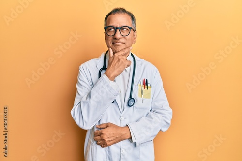 Middle age indian man wearing doctor coat and stethoscope with hand on chin thinking about question, pensive expression. smiling with thoughtful face. doubt concept. © Krakenimages.com