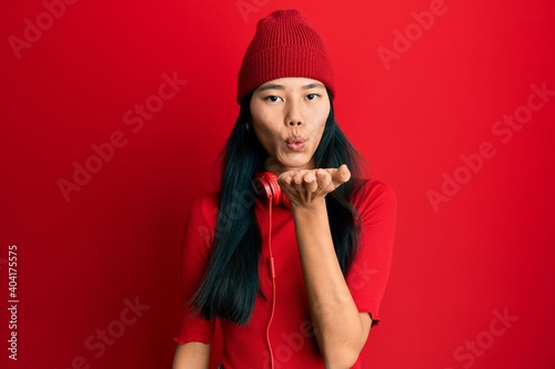 Young chinese woman listening to music using headphones looking at the camera blowing a kiss with hand on air being lovely and sexy. love expression.
