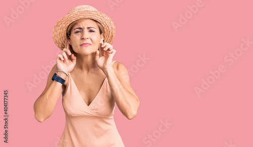 Young blonde woman wearing summer hat trying to hear both hands on ear gesture, curious for gossip. hearing problem, deaf