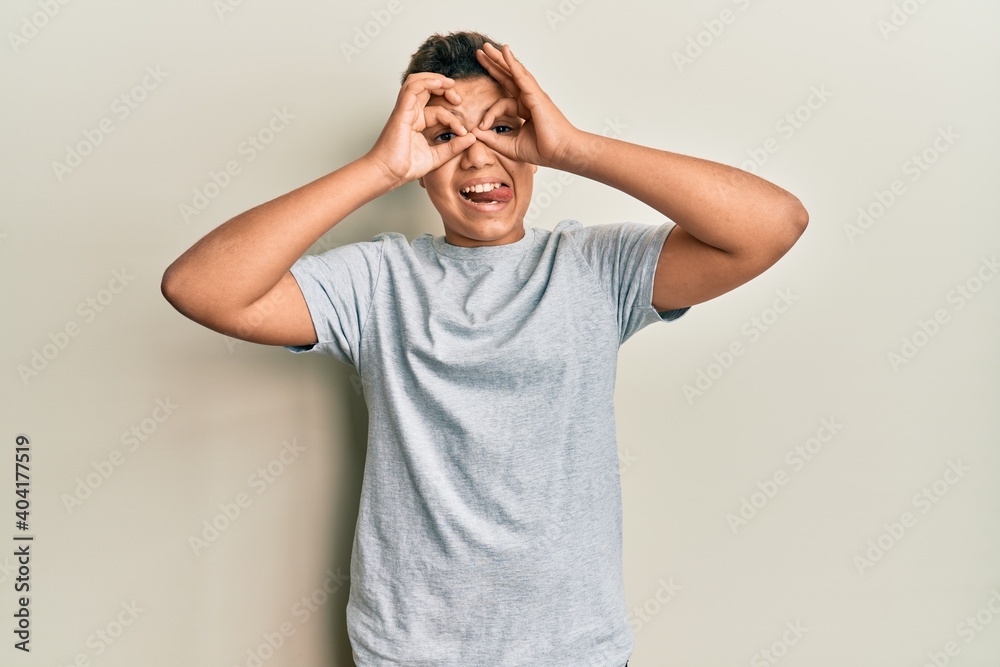 Teenager hispanic boy wearing casual grey t shirt doing ok gesture like binoculars sticking tongue out, eyes looking through fingers. crazy expression.