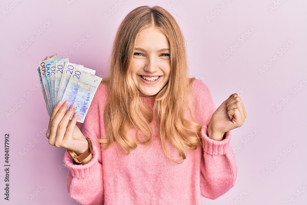 Beautiful young caucasian girl holding swedish krona banknotes screaming proud, celebrating victory and success very excited with raised arm Beautiful young caucasian girl holding swedish krona banknotes screaming proud, celebrating victory and success very excited with raised arm