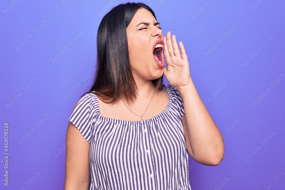Young beautiful brunette woman wearing casual striped t-shirt standing over purple background shouting and screaming loud to side with hand on mouth. Communication concept. Young beautiful brunette woman wearing casual striped t-shirt standing over purple background shouting and screaming loud to side with hand on mouth. Communication concept.
