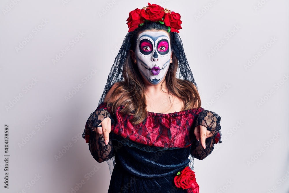 Young woman wearing day of the dead costume over white pointing down with fingers showing advertisement, surprised face and open mouth Young woman wearing day of the dead costume over white pointing down with fingers showing advertisement, surprised face and open mouth