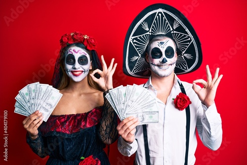 Couple wearing day of the dead costume holding dollars doing ok sign with fingers, smiling friendly gesturing excellent symbol © Krakenimages.com