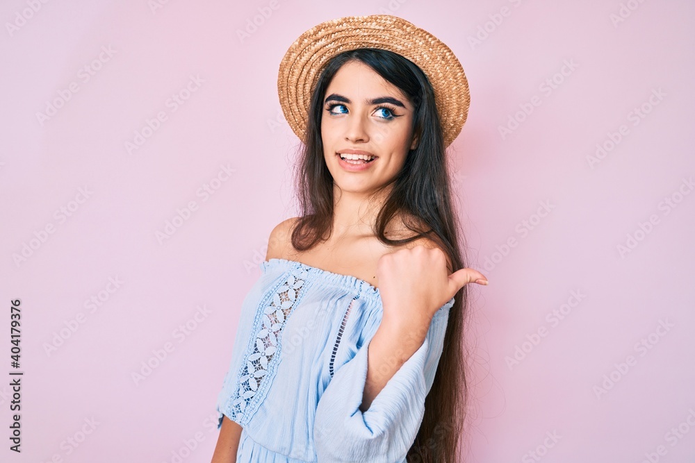 Brunette teenager girl wearing summer hat smiling with happy face looking and pointing to the side with thumb up. Brunette teenager girl wearing summer hat smiling with happy face looking and pointing to the side with thumb up.
