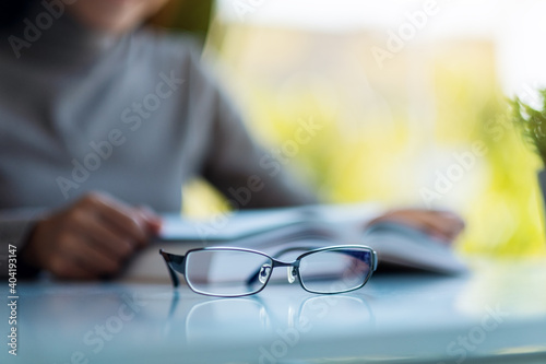 An eye glasses on the table with blurred of a woman reading book in background