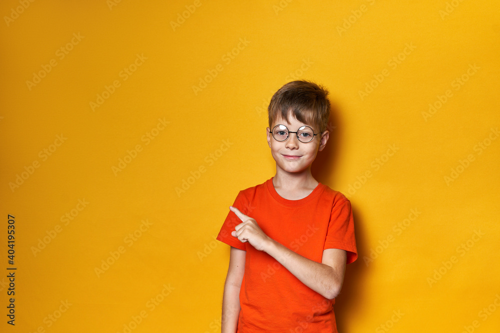 Smiling cute boy in round glasses and orange t-shirt on a yellow isolated background points his finger to the side to the place for text, banner Smiling cute boy in round glasses and orange t-shirt on a yellow isolated background points his finger to the side to the place for text, banner