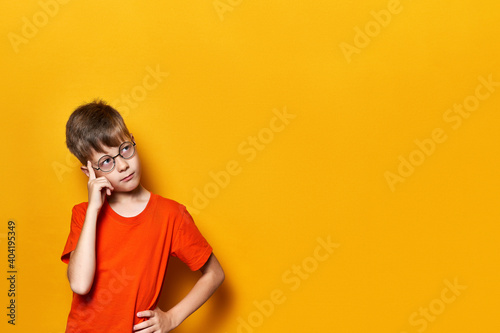 Child in thought Isolated on yellow background in orange t-shirt. Boy wearing glasses with a big Idea