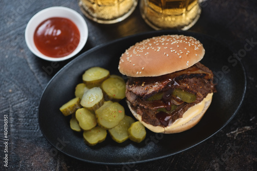 Black plate with brisket sandwich, pickles and beer, horizontal shot on a dark brown stone background