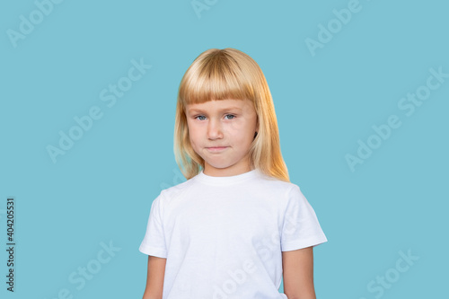 Sarcastic kid. Naughty childhood. Innocent beauty. Portrait of shy confused cute blonde little girl in white t-shirt looking at camera isolated on blue copy space background.