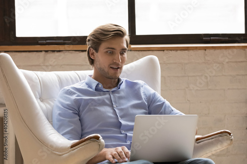 Shocked young Caucasian man sit in chair look at laptop screen stunned by unexpected news online. Amazed male relax in armchair shopping on web on computer surprised by sale discount or deal.