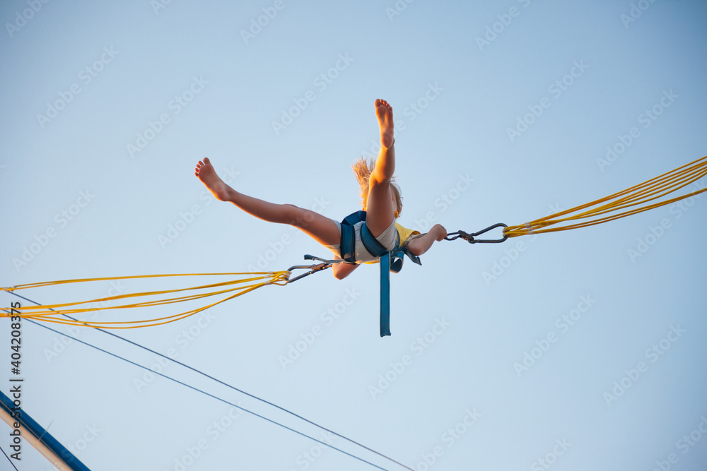 A little cheerful girl flies on springy bright elastic bands and jumps on a trampoline enjoying the long-awaited vacation in the warm sun - obrazy, fototapety, plakaty 