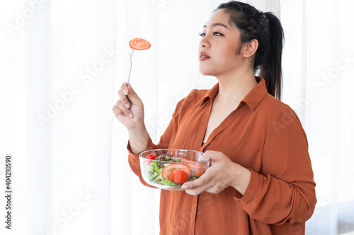 Overweight Woman Eating Bowl Of Fresh Vegetables. Obesity young women eat green organic vegetables. Obese female holding bowl salad vegetarian while eating tomatoes. Overweight woman eating salad.
