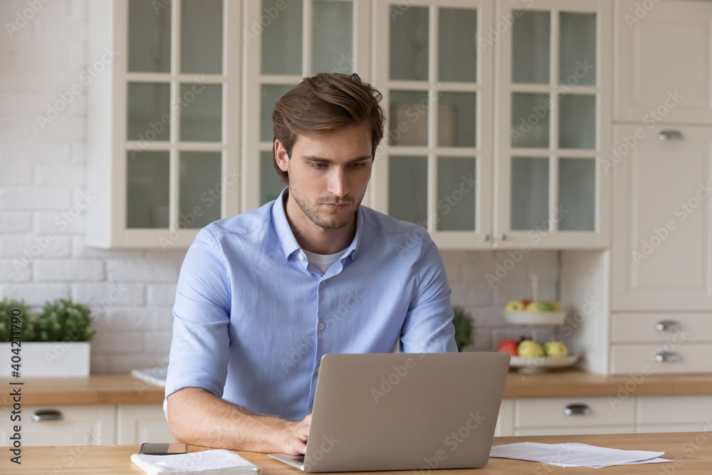 Focused young Caucasian man sit at table at kitchen look at laptop screen browsing internet on device. Serious millennial male work on computer with paperwork documents online at home office. Focused young Caucasian man sit at table at kitchen look at laptop screen browsing internet on device. Serious millennial male work on computer with paperwork documents online at home office.