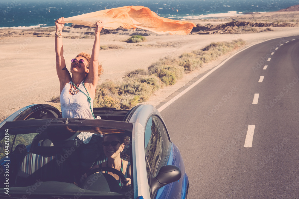 Concept of travel happiness with adult beautiful couple of women driving a car and enjoying the freedom together in friendship - people on the road with convertible vehicle and ocean background