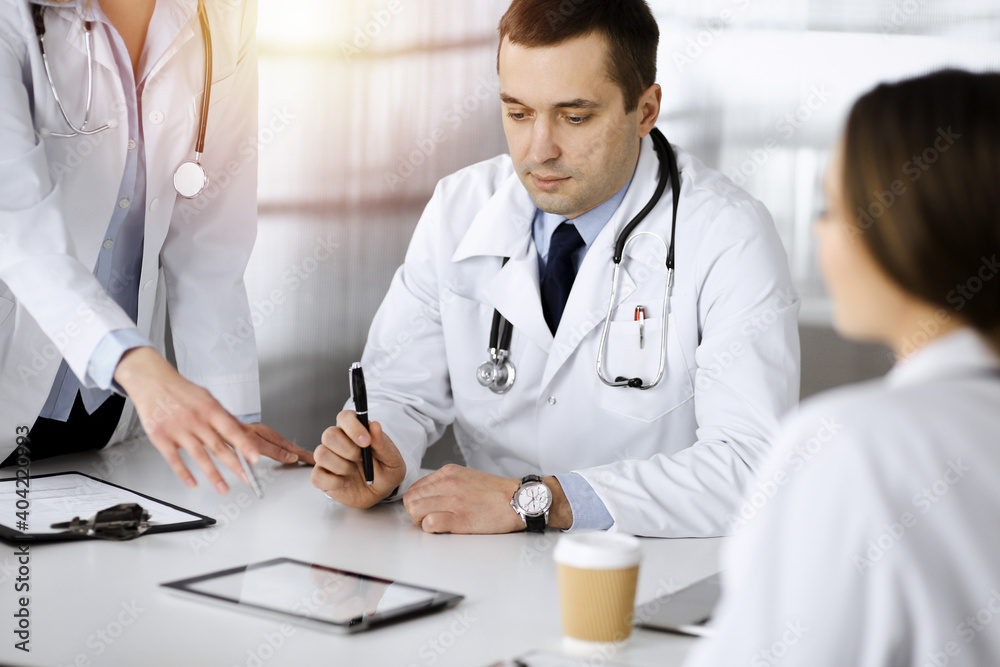 Professional middle aged doctor is talking to his colleagues, while sitting at the desk in a sunny cabinet at hospital. Team of doctors discussing blood sample's results in a clinic. Perfect medicine Professional middle aged doctor is talking to his colleagues, while sitting at the desk in a sunny cabinet at hospital. Team of doctors discussing blood sample's results in a clinic. Perfect medicine