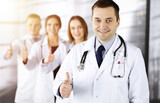 Group of professional doctors are standing as a team with thumbs up in a sunny hospital office, ready to help their patients. Modern medical help, insurance in health care and medicine concept