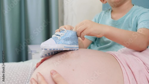 Closeup of older son walking with baby boots acroos big belly of pregnant mother lying on bed at bedroom photo