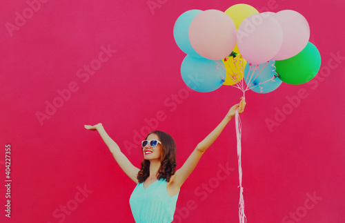 Happy joyful beautiful woman with bunch of colorful balloons raising her hands up on a pink background