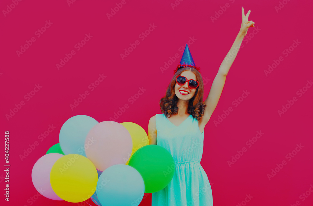 Fototapeta premium Happy surprised smiling young woman in birthday cap with colorful balloons on a pink background