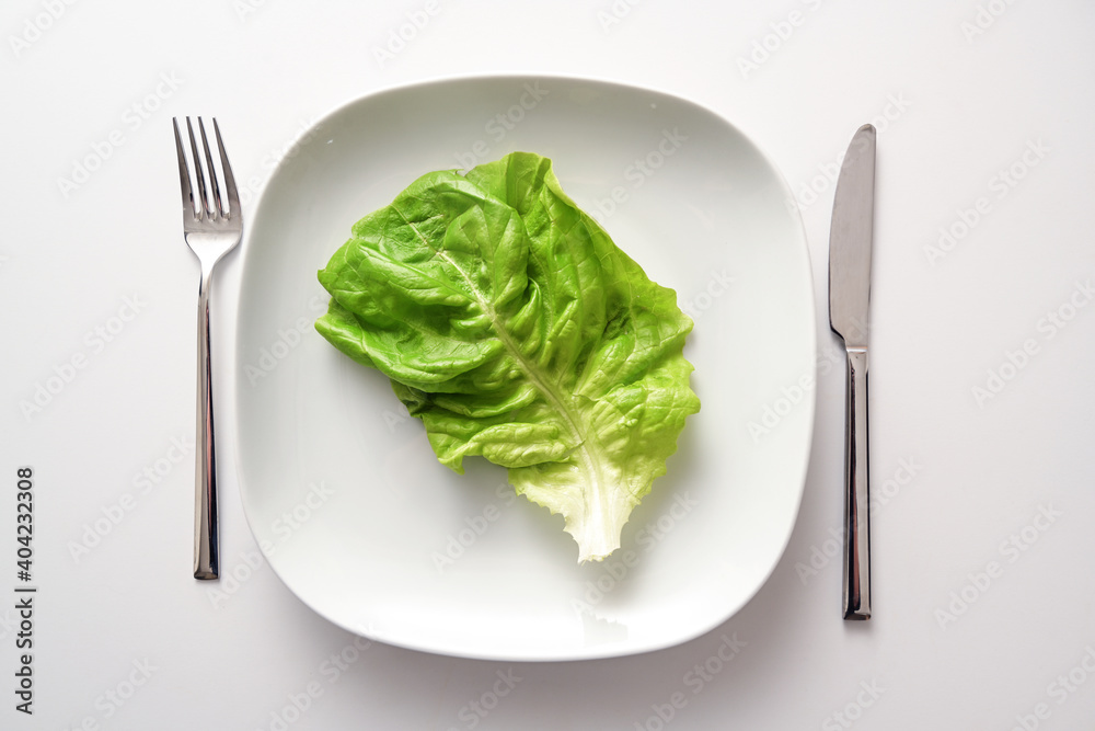 Green lettuce leaf on a square plate and cutlery on a white background, healthy diet to lose weight as a resolution in the new year, copy space, high angle view from above Green lettuce leaf on a square plate and cutlery on a white background, healthy diet to lose weight as a resolution in the new year, copy space, high angle view from above