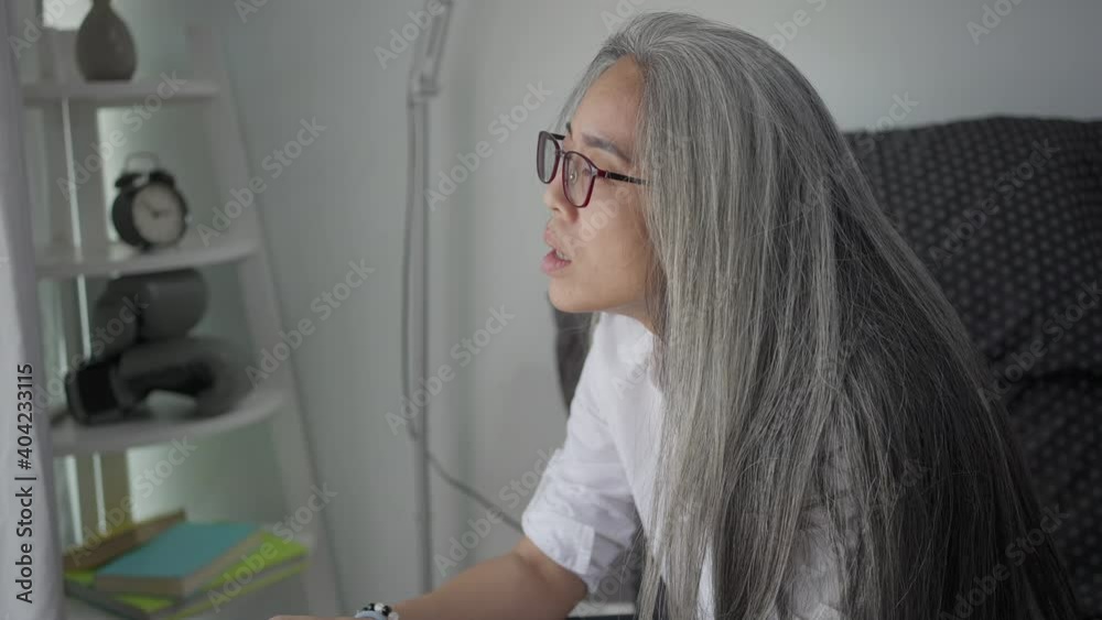 Worried  grey-haired pretty woman in glasses posing sits on the bed at bedroom. Peaceful  grey-haired lady resting and stress looking away thinking. 