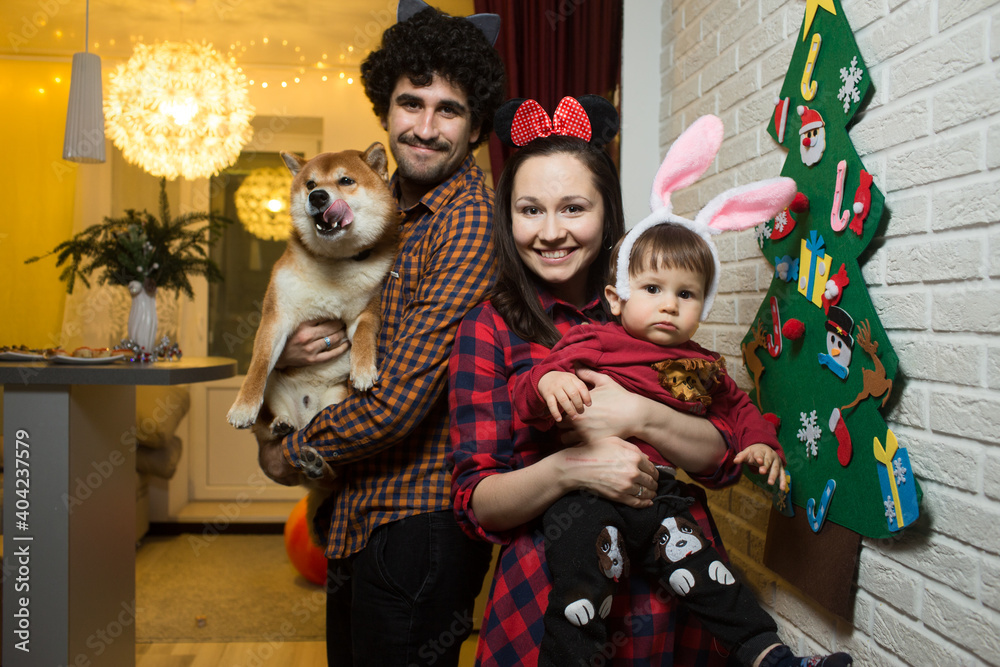 A young family of a man, a woman with a toddler child and a red shiba inu dog, stand nearby and hug. They put on fancy dress and stand by the tree. They celebrate the holiday together