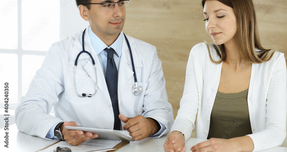 Fototapeta premium Doctor and patient discussing medical exam results while sitting at the desk in clinic. Male physician using tablet computer for filling up medication history record of young woman. Data in medicine
