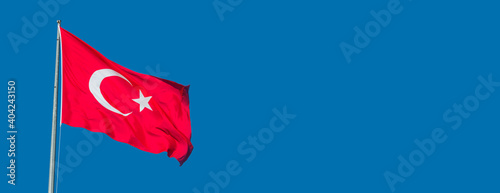 Banner with red Turkish national flag holding white crescent moon and star on the flag at blue sky and copy space for text.