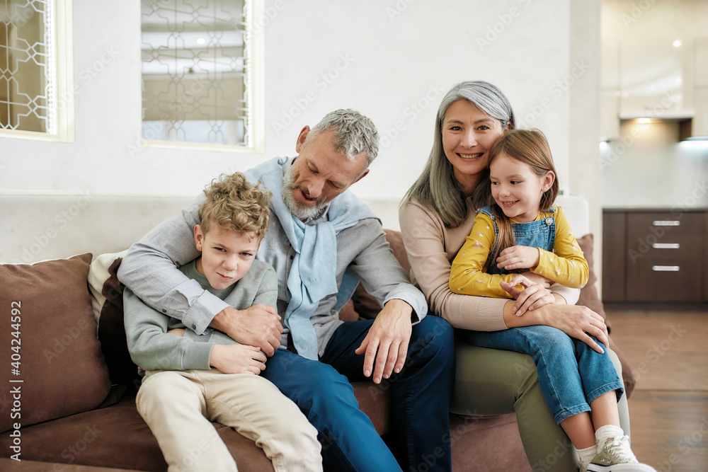 Grandparents with grandchildren hugging and smiling while sitting together on sofa in the living room at home