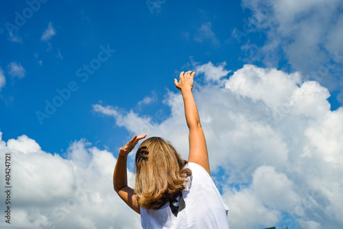 A woman stands with her back to the photographer in a white T-shirt and against a blue sky with clouds. Reaches her hand to the sky. 