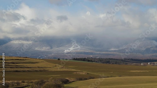 Timelapse of clouds over Lomnicky peak in High Tatras rocky mountains in windy winter day, Slovakia. Landscape of fields and meadows under mountains not covered with snow due to climate changes.