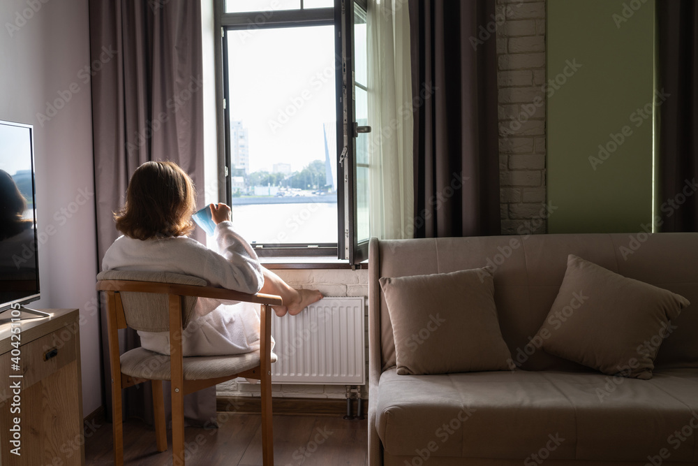 Back view of beautiful young woman in bathrobe with cup of coffee sitting on armchair and enjoying near open window at sunny morning. Back view of beautiful young woman in bathrobe with cup of coffee sitting on armchair and enjoying near open window at sunny morning.