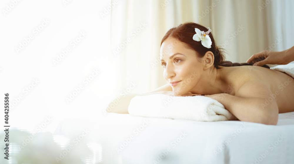 Pretty brunette woman enjoying treatment with hot stones in sunny spa salon. Beauty concept. Cold toned picture Pretty brunette woman enjoying treatment with hot stones in sunny spa salon. Beauty concept. Cold toned picture