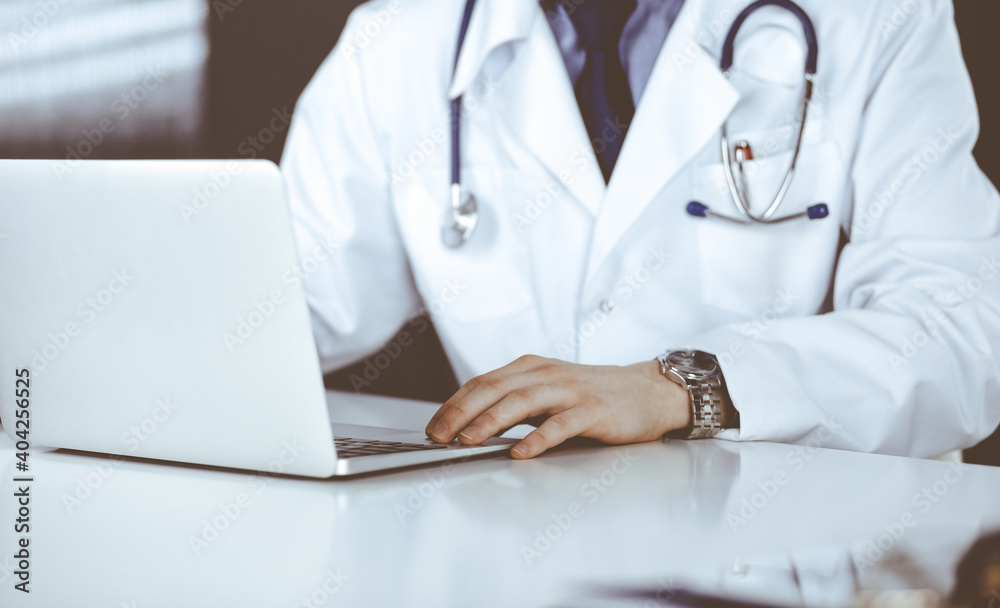 Unknown male doctor sitting and working with laptop in clinic at his working place, close-up. Young physician at work. Perfect medical service, medicine concept Unknown male doctor sitting and working with laptop in clinic at his working place, close-up. Young physician at work. Perfect medical service, medicine concept