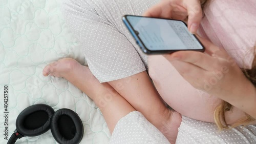 Closeup of pregnant womansitting on bed and typing message on smartphone after listening music in headphones. Concept of early music, art and creativity development photo