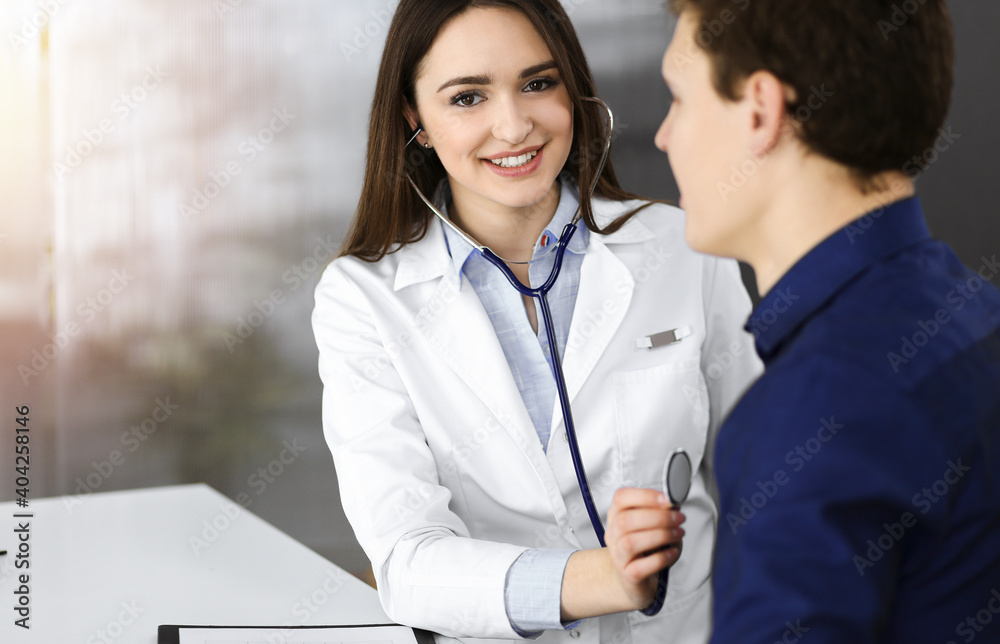 Fototapeta premium Friendly young woman-doctor is listening to her patient's heart with a stethoscope, while they are sitting together at the desk in a cabinet. Physician at work in a hospital. Medicine and healthcare